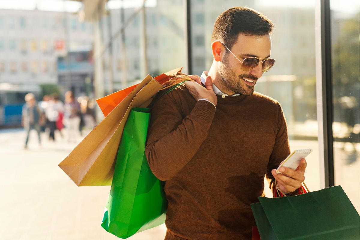 The picture shows a man carrying shopping bags while using a phone which means avoiding lifestyle inflation keeps your financial goals in sight.