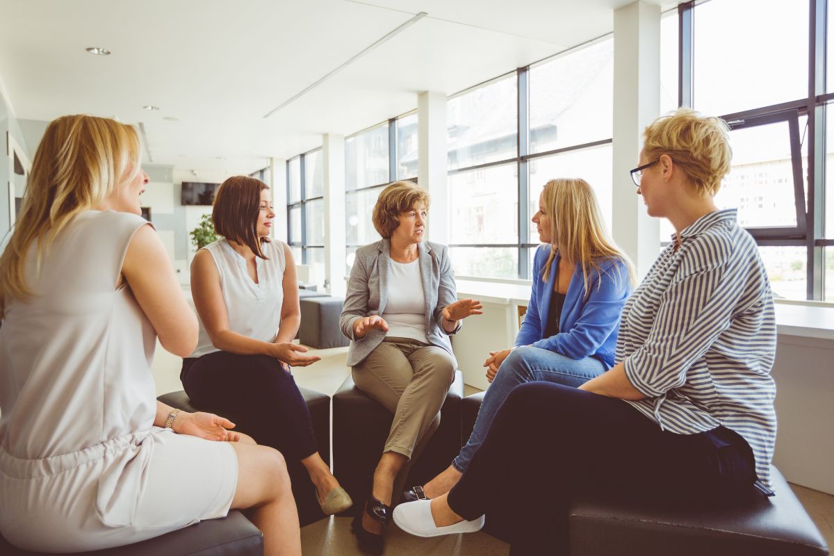 The image shows a group of women talking together in a room referring to surrounding yourself with financially responsible people helps you stay motivated and easier to build wealth with others' support. 