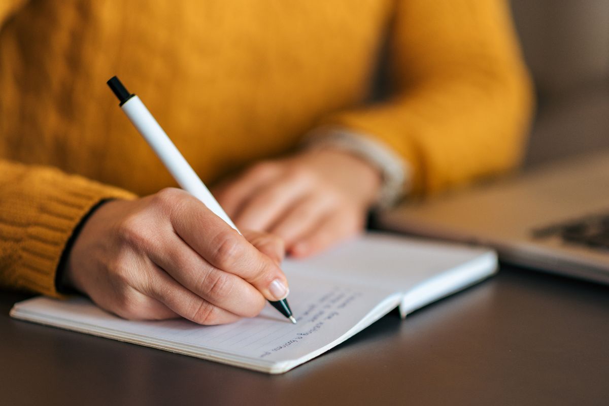 The image shows a laptop and a girl's hand and writing on the notebook referring to writing a letter to your future financially independent self as a great way to stay focused on becoming a millionaire or financially stable.