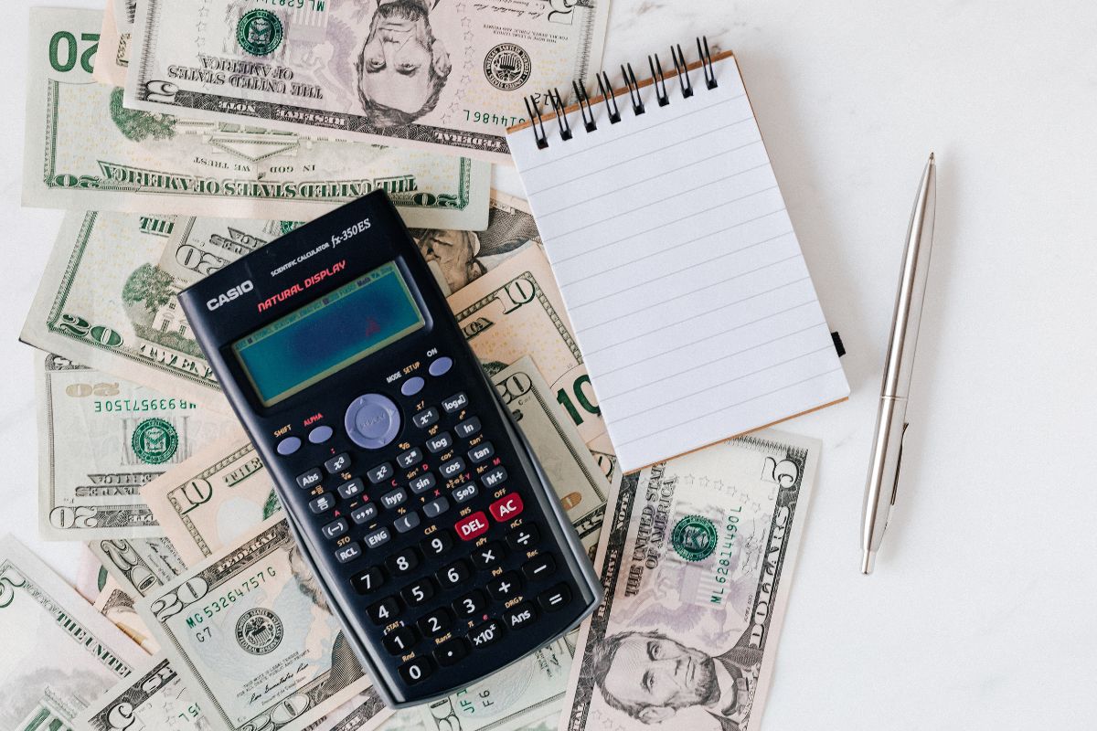 The image shows a pen, a mini notebook, a calculator, and dollar bills to assess your current financial situation helps you figure out if you're on track or need to make changes.