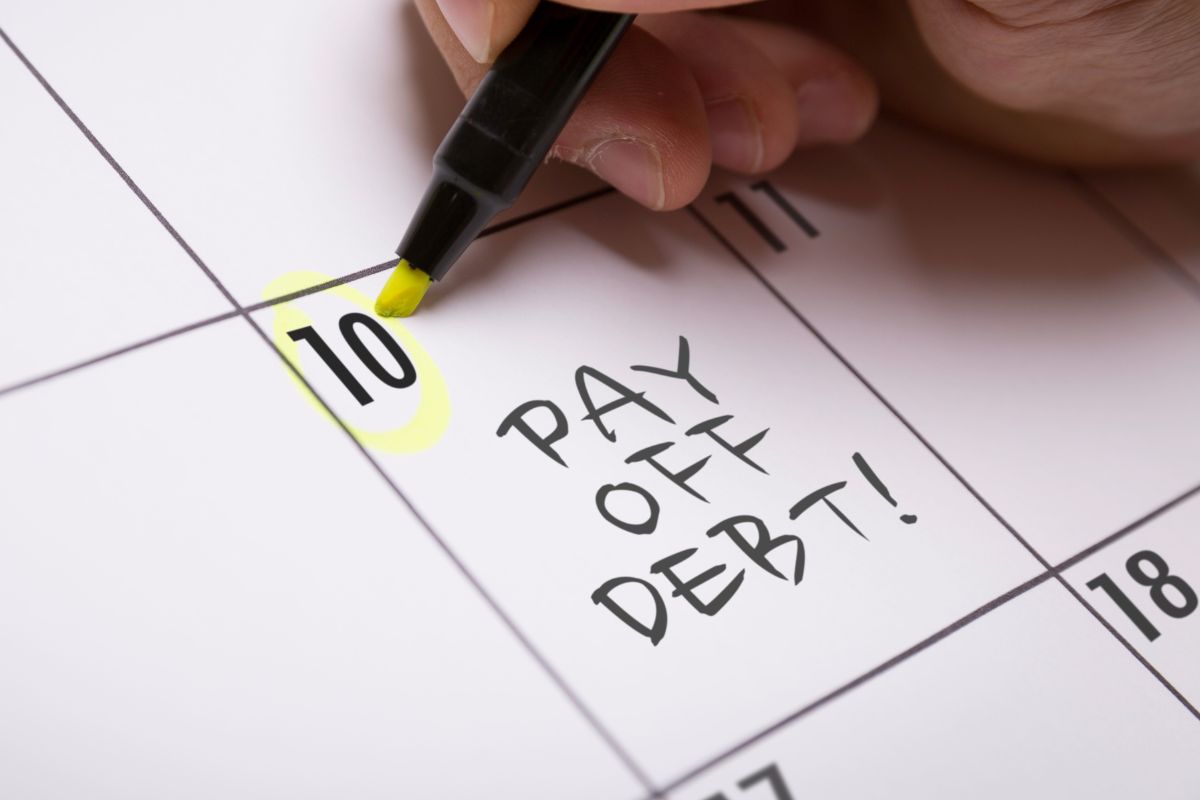 The image shows a man's hand writing pay off debt on a calendar.