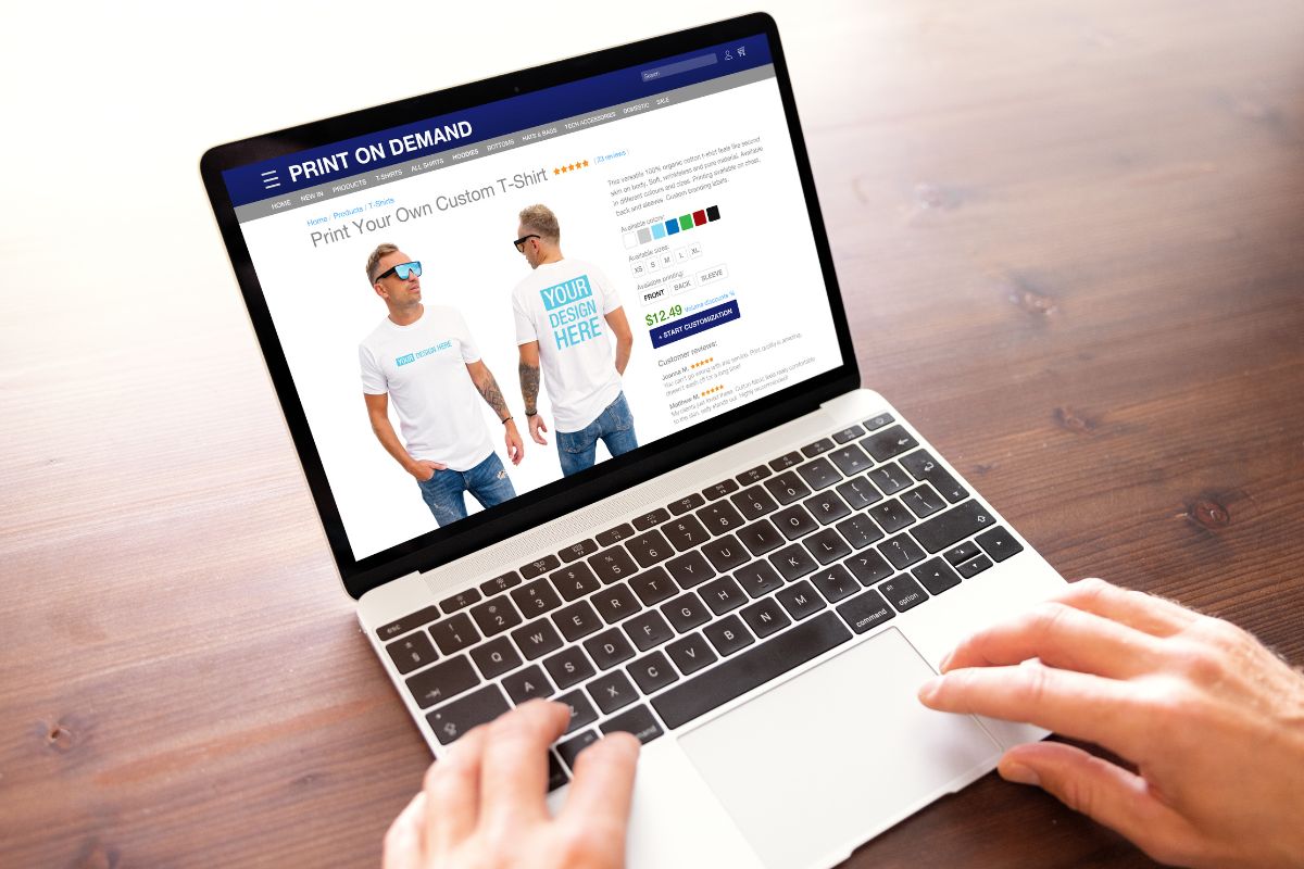 The picture shows a man's hand using a laptop for print-on-demand designing.