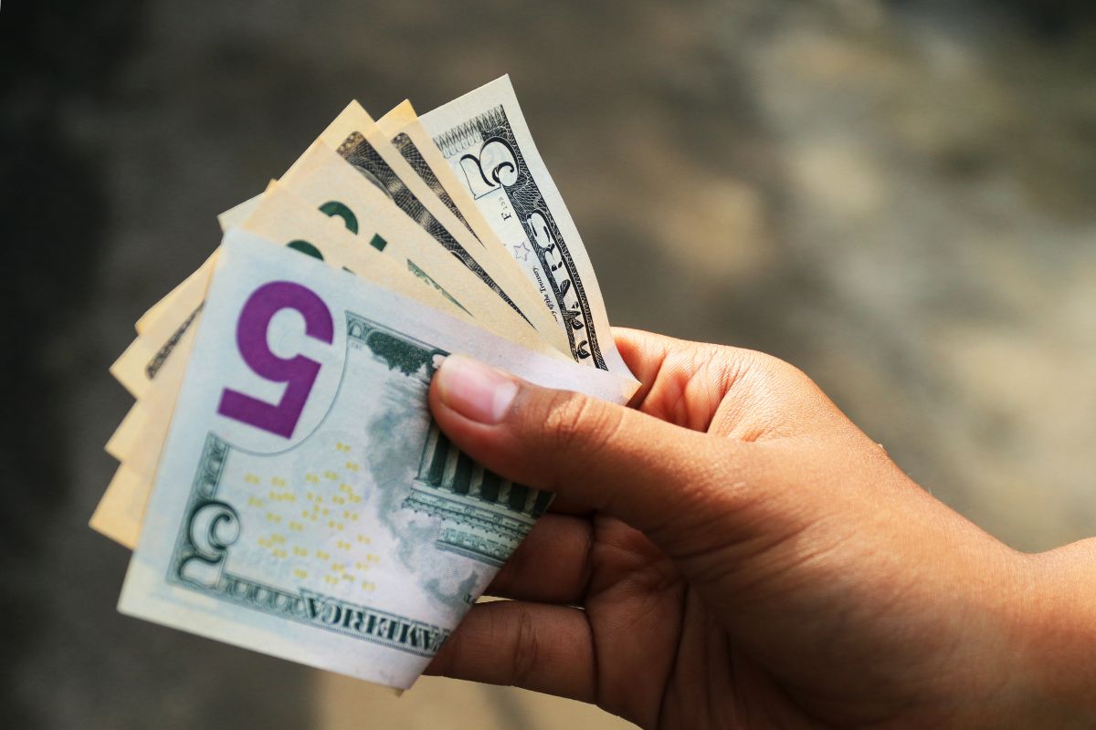 The image shows a woman's hand holding $5 bills.