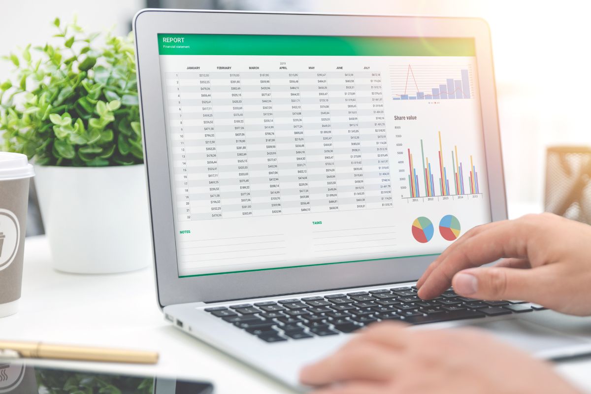 The image shows a man's hand using a laptop with a budget spreadsheet.
