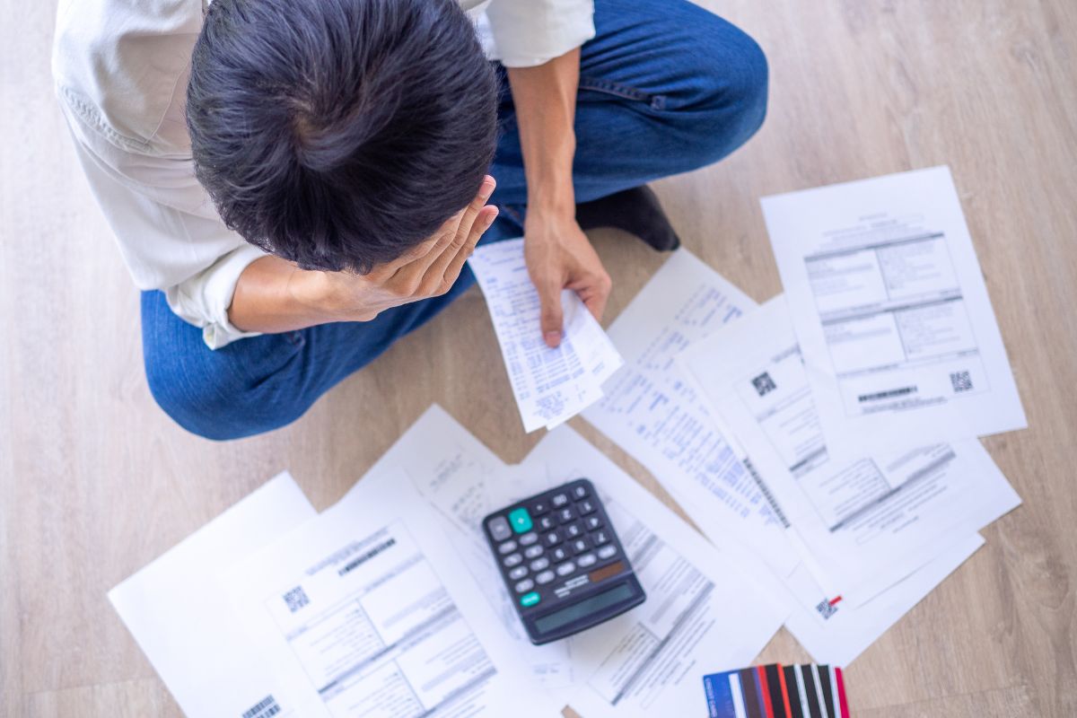 The image shows papers on the floor, a calculator, and a frustrated man holding receipts referring to stop making these money mistakes and take control of your finances with these steps.
