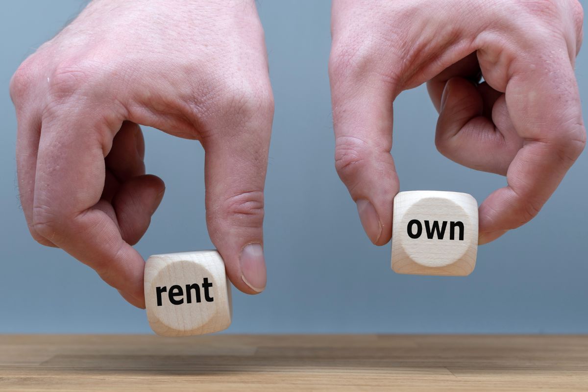 The image shows a man's hand holding wooden cubes with notes referring to rent vs. own is a good example of financial stability differences.