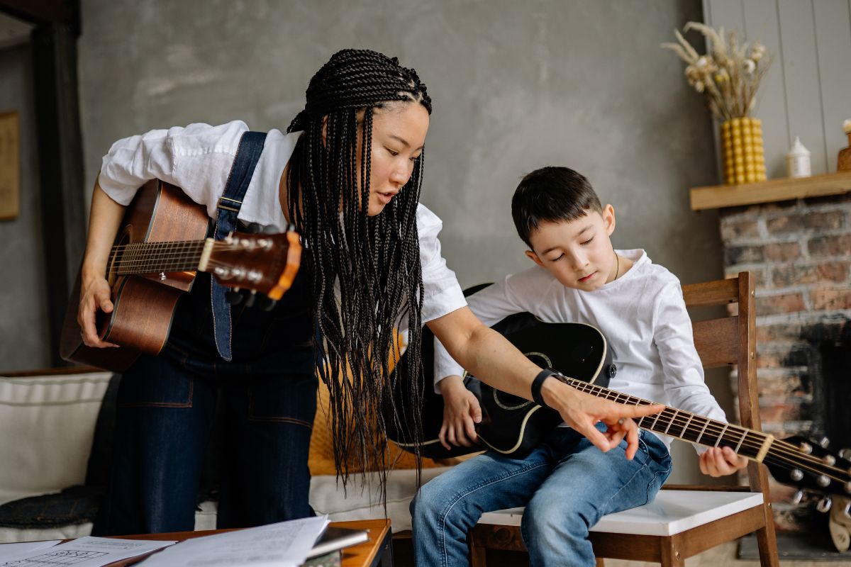 A picture of a lady teaching a kid how to play the guitar.