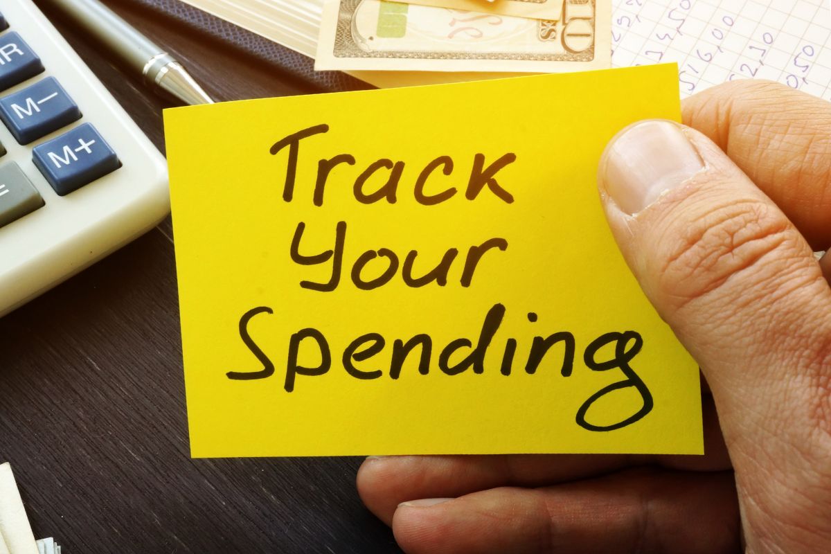 The picture is about a man's hand holding a paper with Track Your Spending notes.