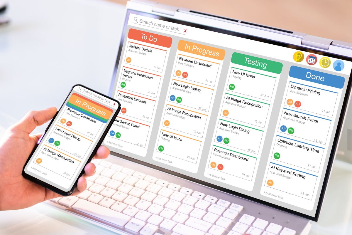 The picture shows a man's hand using a phone and a laptop for project management.