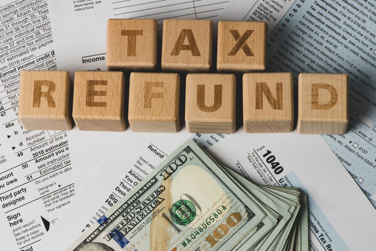 The picture shows papers with notes, wooden cubes with letters, and dollar bills referring to spending tax refunds vs investing it is an easy money life hack that a lot of people overlook. 