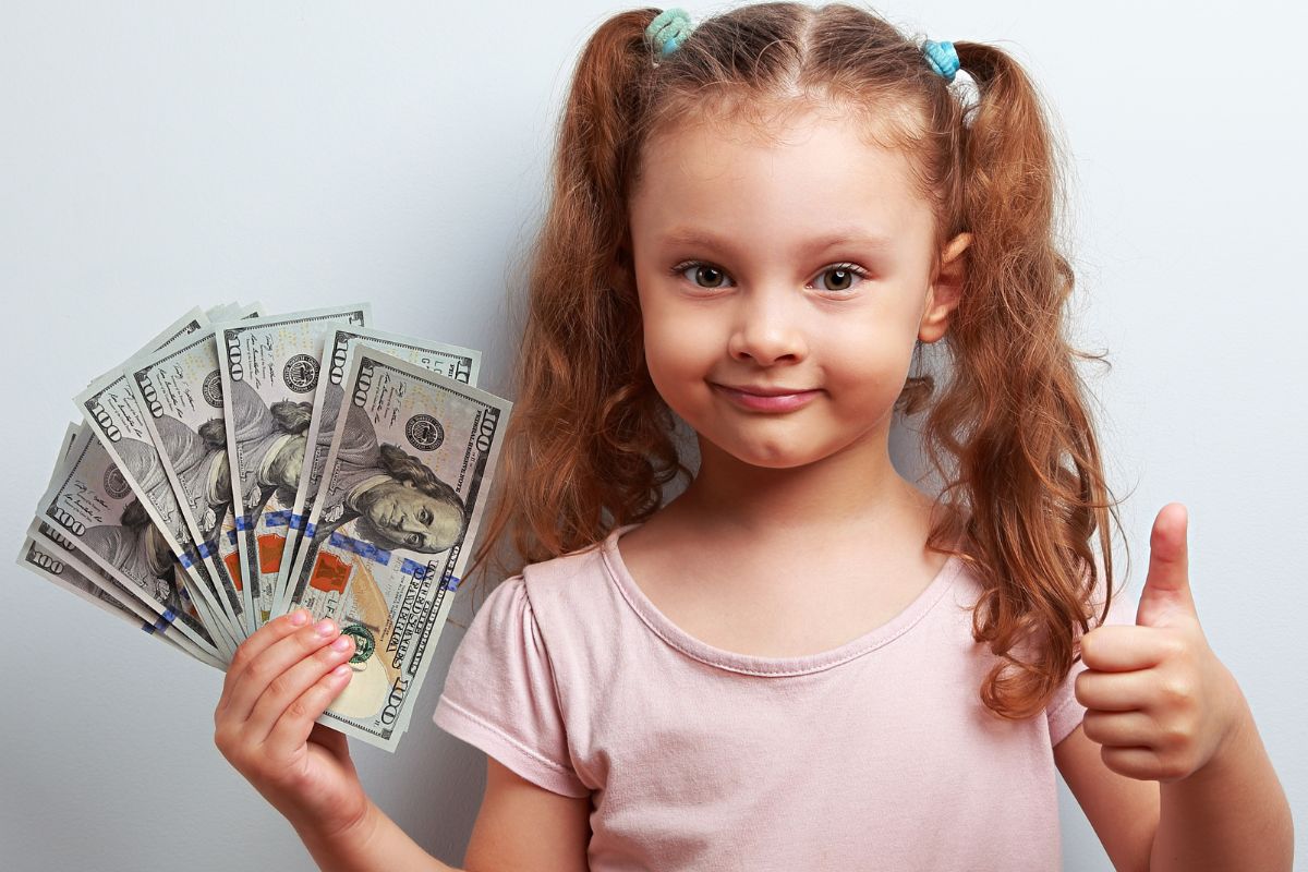 The photo of a kid holding dollar bills while doing an ok sign referring to teaching kids about money.