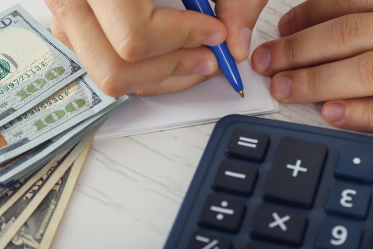 The picture shows a calculator, dollar bills, and a man's hand and writing on the paper which means focusing on mastering one money habit at a time.