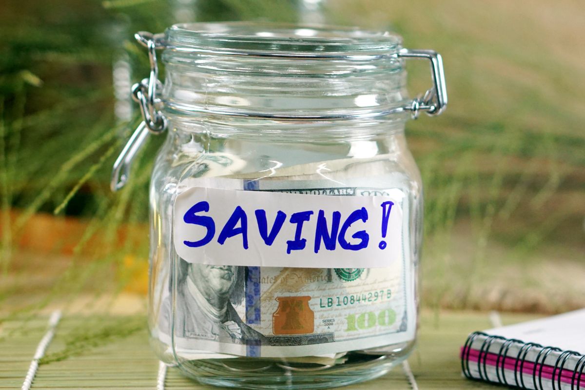 The image shows a notebook and a jar filled with dollar bills and a savings note.