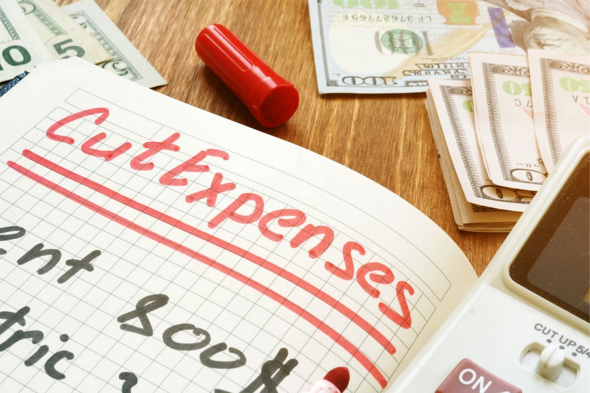The image of a notebook with cut expenses wording and dollar bills.