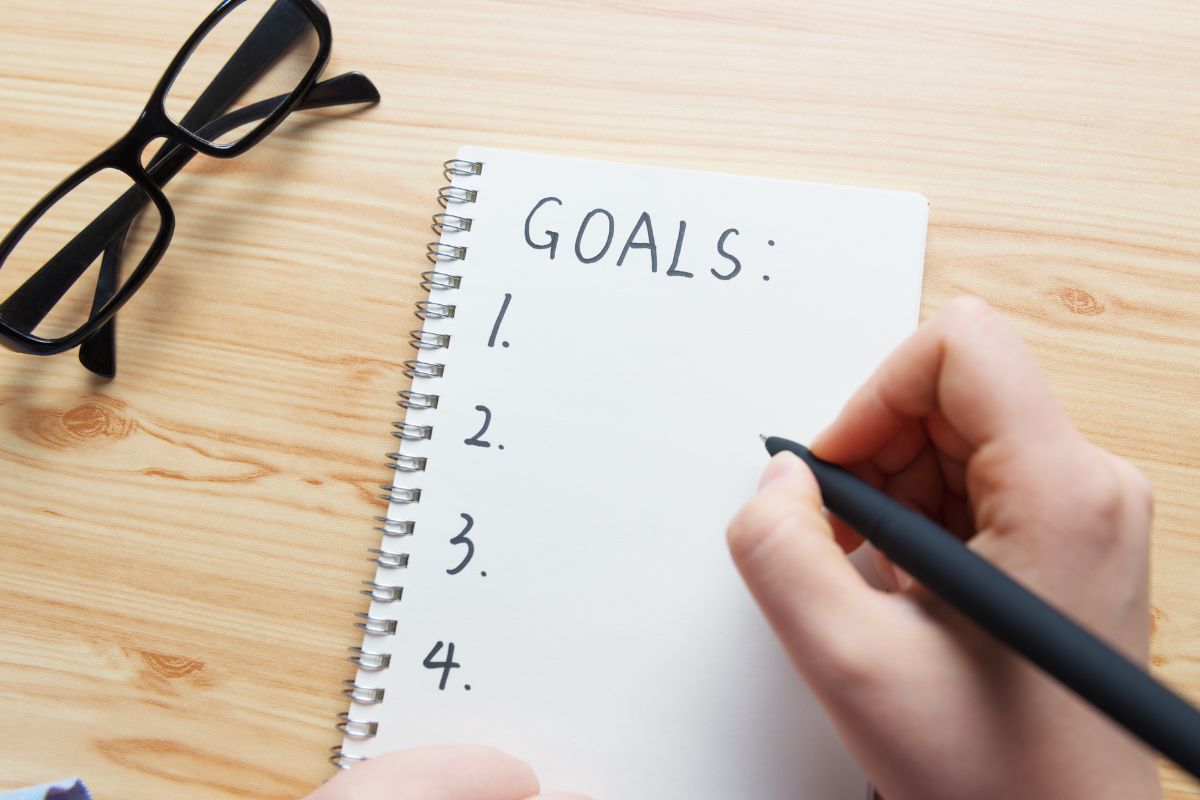 An image shows a lady writing her goals in a notebook.