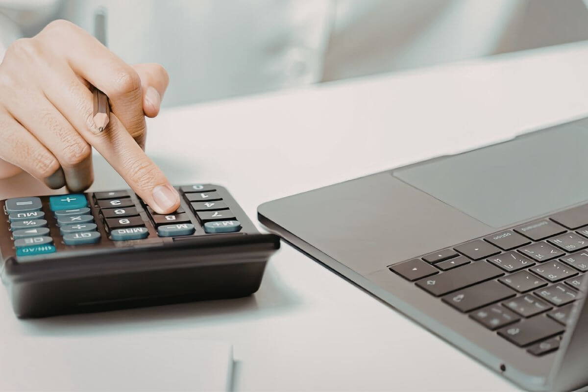 A imagem mostra a mão de uma mulher usando uma calculadora e um laptop.