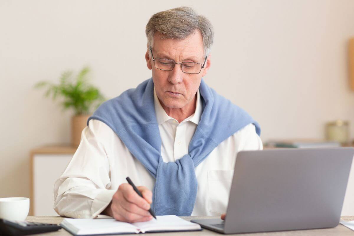 A imagem de um homem maduro escrevendo algo enquanto usa um laptop.